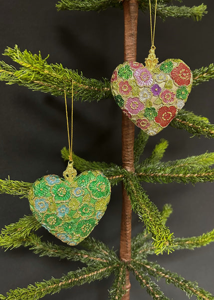 Two heart-shaped ornaments with textured floral details, hanging from a tree. One ornament is green with multicolor floral patterns, and the other is multicolor with a floral pattern on a green background.