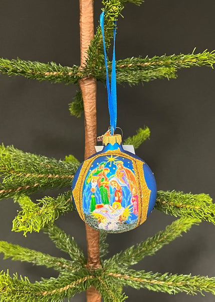 A hand-painted, mouth-blown glass ball Christmas ornament with a nativity scene design, hanging from a blue ribbon on a Christmas tree.