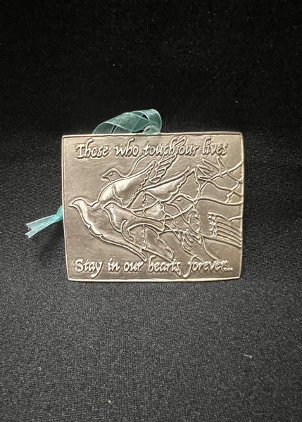 A pewter remembrance plaque with a bird design and an inscription that reads 'There who touched our lives, stay in our hearts forever.' It includes a ribbon for hanging and is presented against a black background.