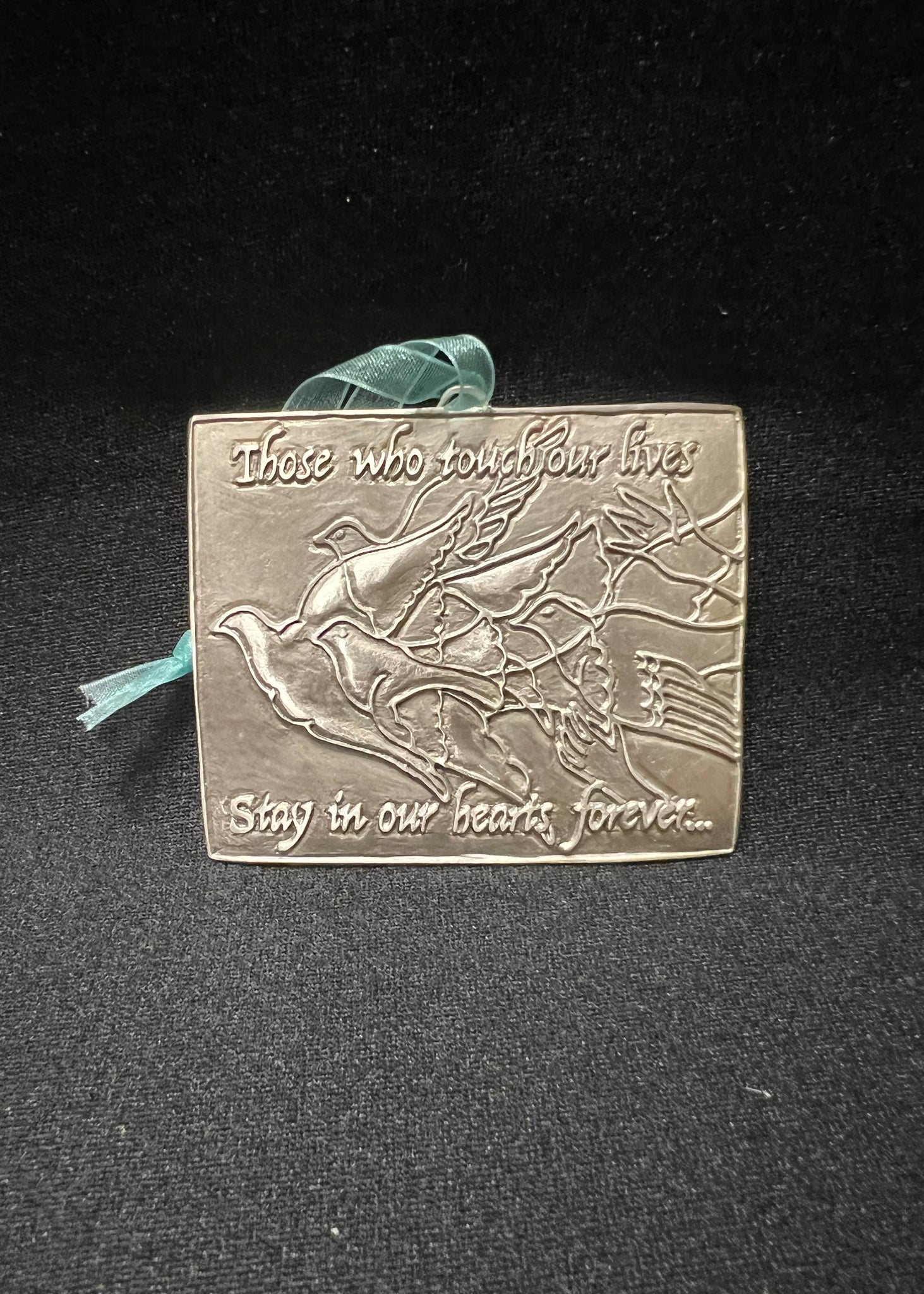 A pewter remembrance plaque with a bird design and an inscription that reads 'There who touched our lives, stay in our hearts forever.' It includes a ribbon for hanging and is presented against a black background.