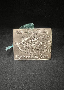 A pewter remembrance plaque with a bird design and an inscription that reads 'There who touched our lives, stay in our hearts forever.' It includes a ribbon for hanging and is presented against a black background.